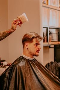 Side view of young bearded man sitting in cape in barbershop while crop anonymous tattooed stylist using brush to wipe hair